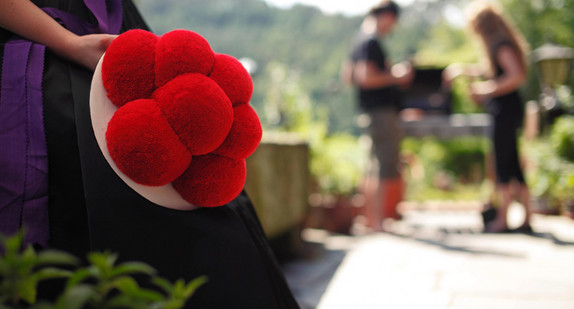 Eine Person in traditioneller Tracht mit einem Bollenhut, dessen rote Wollbollen gut sichtbar im Vordergrund sind. Im unscharfen Hintergrund befinden sich zwei Personen bei einem Grill im Freien, umgeben von grüner Natur.