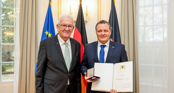 Ministerpräsident Winfried Kretschmann (links) und Thomas Speidel (rechts) mit der Staufermedaille in Gold und der Urkunde. Im Hintergrund stehen Fahnen.