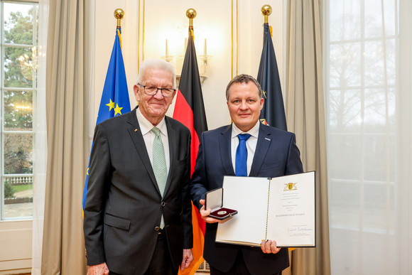 Ministerpräsident Winfried Kretschmann (links) und Thomas Speidel (rechts) mit der Staufermedaille in Gold und der Urkunde. Im Hintergrund stehen Fahnen.