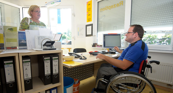 Ein Mann im Rollstuhl arbeitet an einer Rezeption (Quelle: dpa).