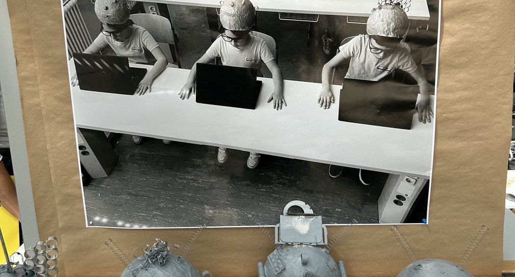 Drei Kinder mit futuristischen Hüten sitzen vor Laptops im Klassenzimmer