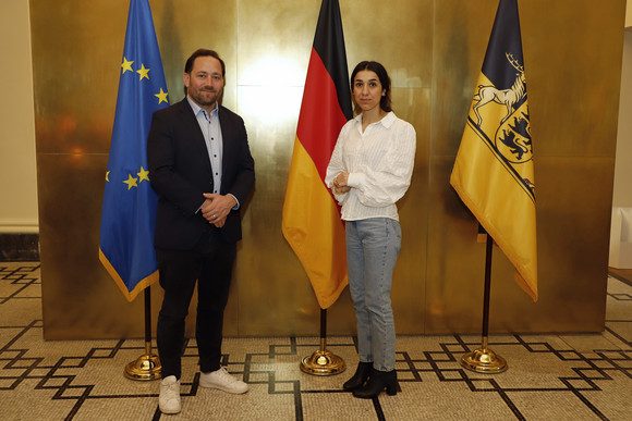 Staatssekretär Florian Hassler (links) und Friedensnobelpreisträgerin Nadia Murad (rechts) stehen in der Villa Reitzenstein in Stuttgart vor Fahnen.