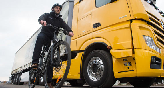 Ein Radfahrer steht mit seinem Fahrrad in der Versuchsabteilung von Mercedes Benz neben einem LKW mit einem Abbiegeassistenten. (Bild:© dpa)