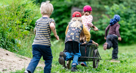 Von hinten sind Kinder zu sehen, die im Wald mit einem Anhänger spielen.