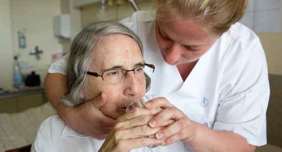 Mit Hilfe einer Schwester führt eine 80-jährige Frau ein Glas Wasser zum Mund und trinkt. 