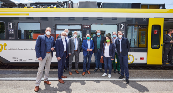 Eine Gruppe mehrerer Menschen steht am Bahnhof vor einem Zug zu einem Gruppenbild zusammen. 