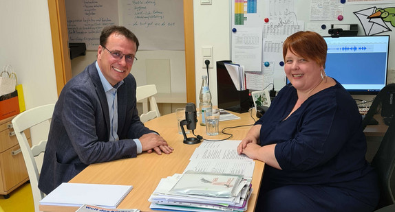Staatssekretär Volker Schebesta und Kita-Leiterin Saskia Franz an einem Schreibtisch, auf dem ein Mikrofon steht