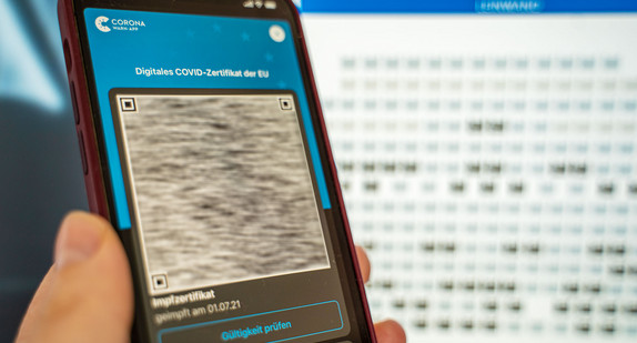 Auf einem Mobiltelefon im Vordergrund ist ein digitales Impfzertifikat vor einem Bildschirm mit einem Kartenbuchungssystem eines Kinos zu sehen. 