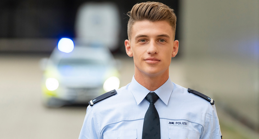 Junger Polizist der Polizei Baden-Württemberg vor Streifenwagen.