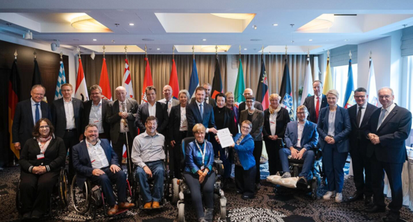 Mitglieder der Jahreskonferenz der Ministerpräsidentinnen und Ministerpräsidenten und die Beauftragten der Länder und des Bundes für die Belange von Menschen mit Behinderungen beim Gruppenbild in Leipzig. Im Hintergrund sieht man die Landesflaggen aller Bundesländer.