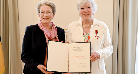 Zwei ältere Frauen, beide halten eine Mappe mit einer Urkunde, die andere trägt eine Auszeichnung am Revers.