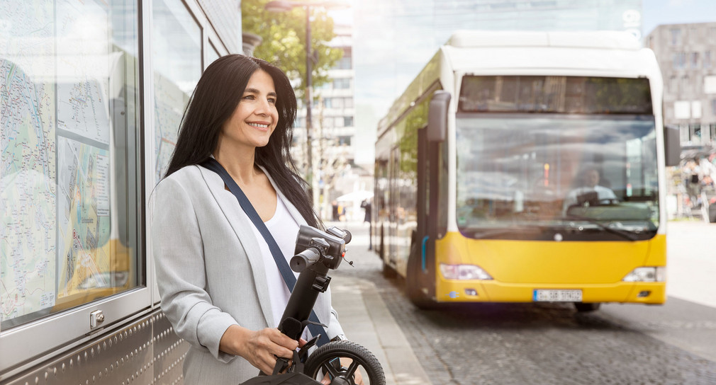 Frau wartet an Haltestelle auf einen hereinfahrenden gelben Bus.