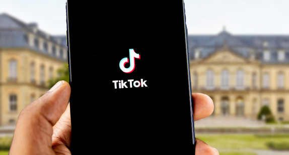 Hand mit Smartphone in der Hand, auf dem Bildschirm sieht man die App TikTok, im Hintergrund ist das Neue Schloss Stuttgart zu sehen