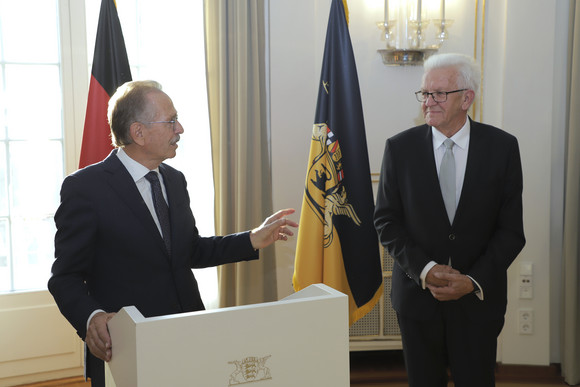 Franz Fehrenbach (links) und Ministerpräsident Winfried Kretschmann (rechts) stehen stehen im Runden Saal der Villa Reitzenstein vor Fahnen.