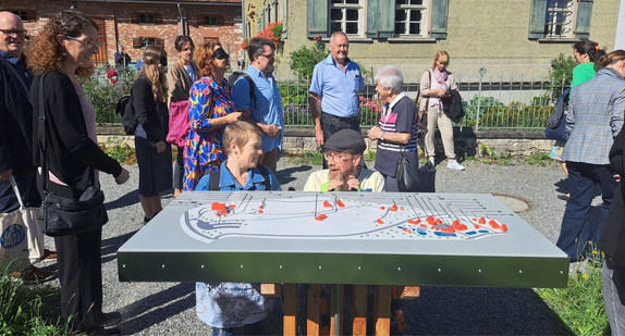 Simone Fischer im Gespräch mit Manfred Klawitter vor einem Tastmodell sowie weitere Teilnehmende des Rundgangs teils mit Sichtbrillen, die eine Sehbehinderung simulieren, auf dem Gelände des Bauernhaus-Museums Wolfegg 