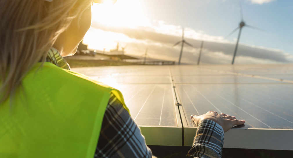 Eine Technikerin steht an einer Freiflächensolaranlage. Im Hintergrund sind mehrere Windkraftanlagen zu sehen.