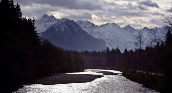 Unter einem dunklen Wolkenhimmel liegen die schneebedeckten Alpen, während im Vordergrund das Wasser der Iller im Gegenlicht glitzert. (Foto: © dpa)