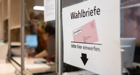 Ein Schild weist in einem Wahlbüro auf eine Wahlurne für die Briefwahl hin.