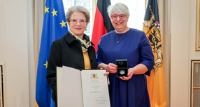 Staatsrätin Barbara Bosch (links) überreicht Irene Armbruster (rechts) die Staufermedaille und eine Urkunde. Im Hintergrdund stehen Fahnen.