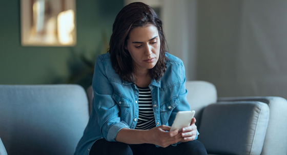 Eine junge Frau sitzt auch einem Sofa und schaut traurig auf ein Smartphone in ihrer Hand.